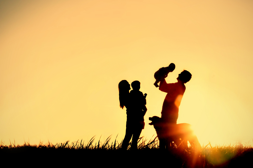 A silhouette of a happy family of four people, mother, father, baby, and child, and their dog in front of a sunsetting sky, with room for copy space or text